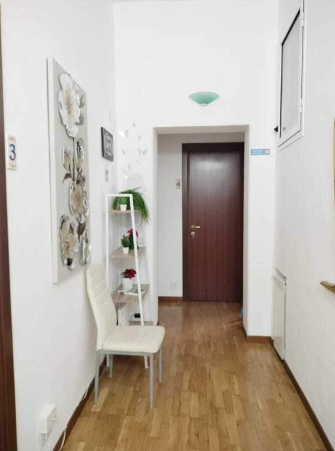 Un corridoio stretto con pavimento in legno, una sedia bianca, uno scaffale bianco con piante e una porta in legno scuro.