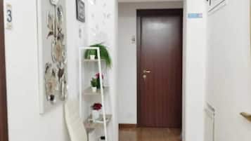 Un corridoio stretto con pavimento in legno, una sedia bianca, uno scaffale bianco con piante e una porta in legno scuro.