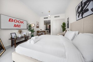 A hotel room with a large bed, a desk, a chair, a dining table, and a kitchen area visible through an open door. - Stayshort - Cozy Studio in Damac Hills That Sleeps 2 (Dubai)