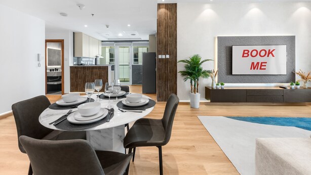 A modern dining area with a round table set for four, a kitchen in the background, and a 'BOOK ME' sign on the wall. - Stayshort - Modern 2BR Apartment in Sulafa Tower Dubai Marina Near to Beach JBR - Sleeps 6 (Dubai)