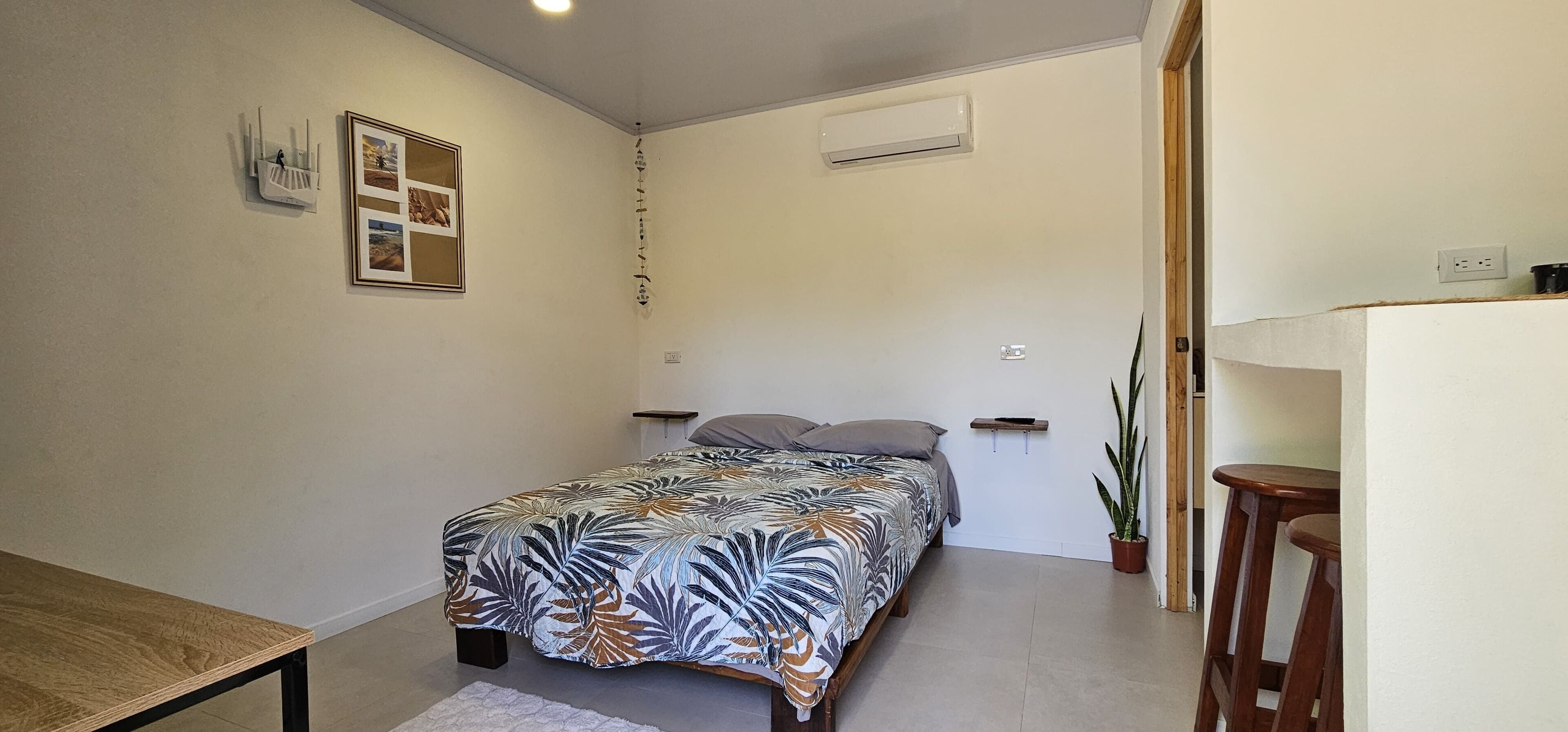 A small, well-lit bedroom with a bed, a bedside table, a wall-mounted air conditioner, and a framed picture on the wall.