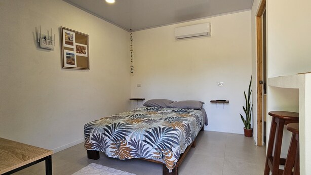 A small, well-lit bedroom with a bed, a bedside table, a wall-mounted air conditioner, and a framed picture on the wall. - Casita Ola Negra, just minutes from the beach—a cozy private studio for 2 people (Puerto Viejo de Talamanca)