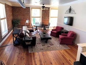 A living room with a brick wall, wooden flooring, a TV mounted on the wall, a dining table with chairs, and a red sofa. - Downtown Wake Forest Loft #1 (Wake Forest)