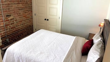 A bedroom with a brick wall, a white door, a bed with white bedding, and a nightstand with a lamp.