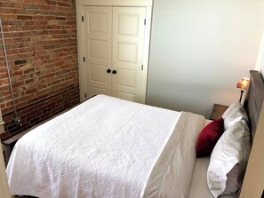 A bedroom with a brick wall, a white door, a bed with white bedding, and a nightstand with a lamp.