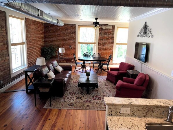 A living room with a brick wall, wooden flooring, a dining table with chairs, a sofa, a red armchair, and a TV mounted on the wall.