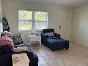 Uma sala de estar com sofá, pufe e mesa lateral com abajur.
