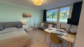A room with a bed, a sofa, a dining table with chairs, a TV, and a window with a view of trees. - Stylish apartment in Munich right next to Südpark (München)