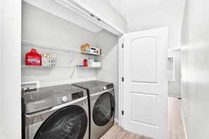 Une buanderie équipée de deux machines à laver Samsung, d’une étagère contenant des produits de nettoyage et d’une porte blanche.