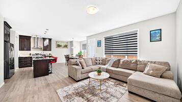 Un salon moderne avec un canapé d’angle, une table basse et un coin cuisine doté de meubles sombres et d’un îlot central.