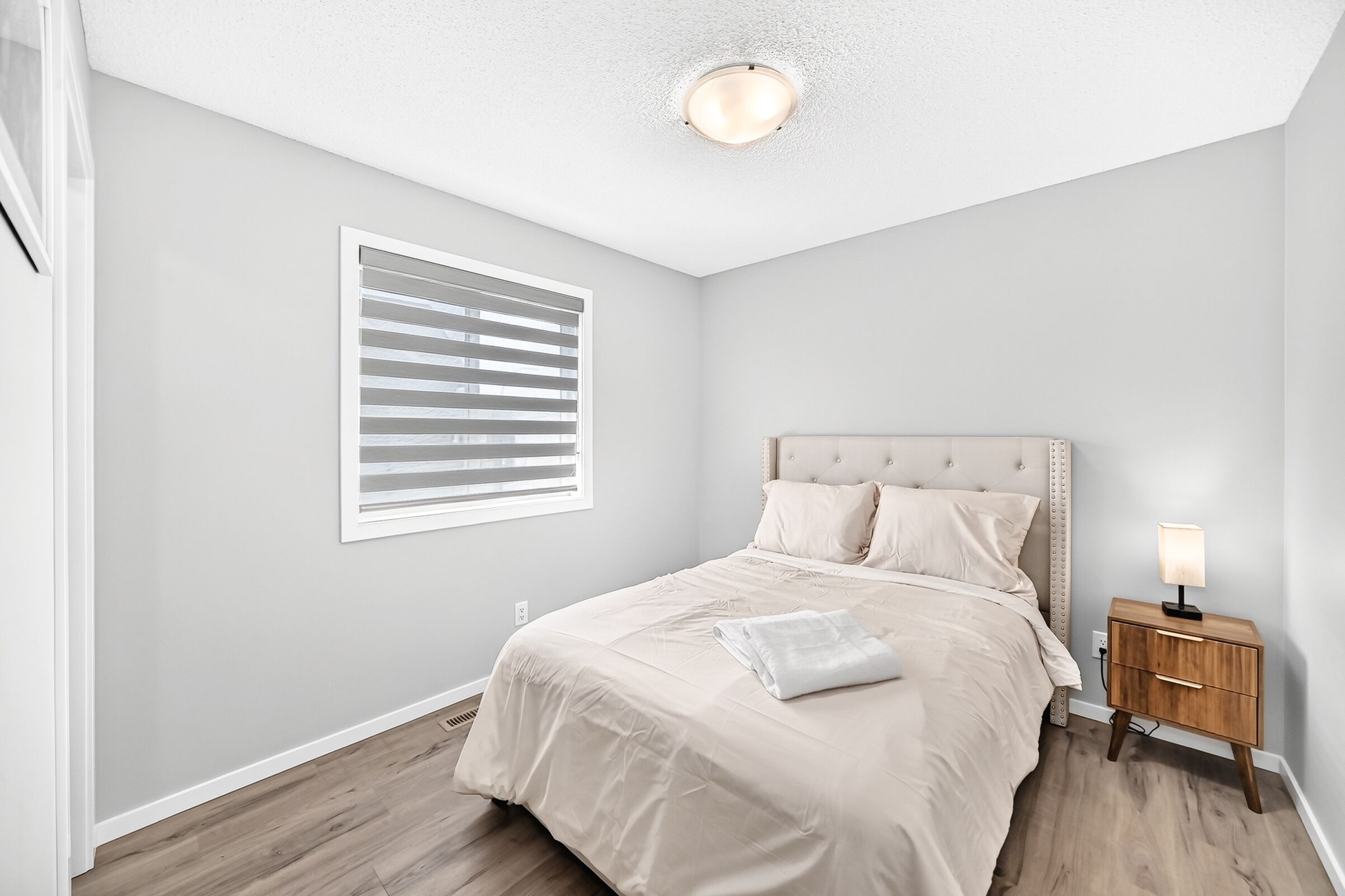 A bedroom with a bed, a nightstand, a lamp, a window with blinds, and a ceiling light.
