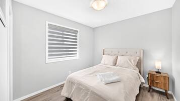 A bedroom with a bed, a nightstand, a lamp, a window with blinds, and a ceiling light.