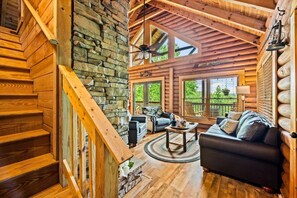 A log cabin interior with a stone wall, wooden staircase, a living area with a sofa and coffee table, and a large window. - Cabin w/ Hot Tub · Hear the Dollywood Whistle (Sevierville)
