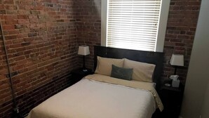 Une chambre avec un mur en briques, un lit avec une tête de lit, deux tables de chevet avec des lampes et une fenêtre avec des stores.