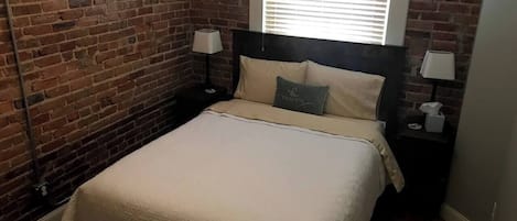 Une chambre avec un mur en briques, un lit avec une tête de lit, deux tables de chevet avec des lampes et une fenêtre avec des stores.
