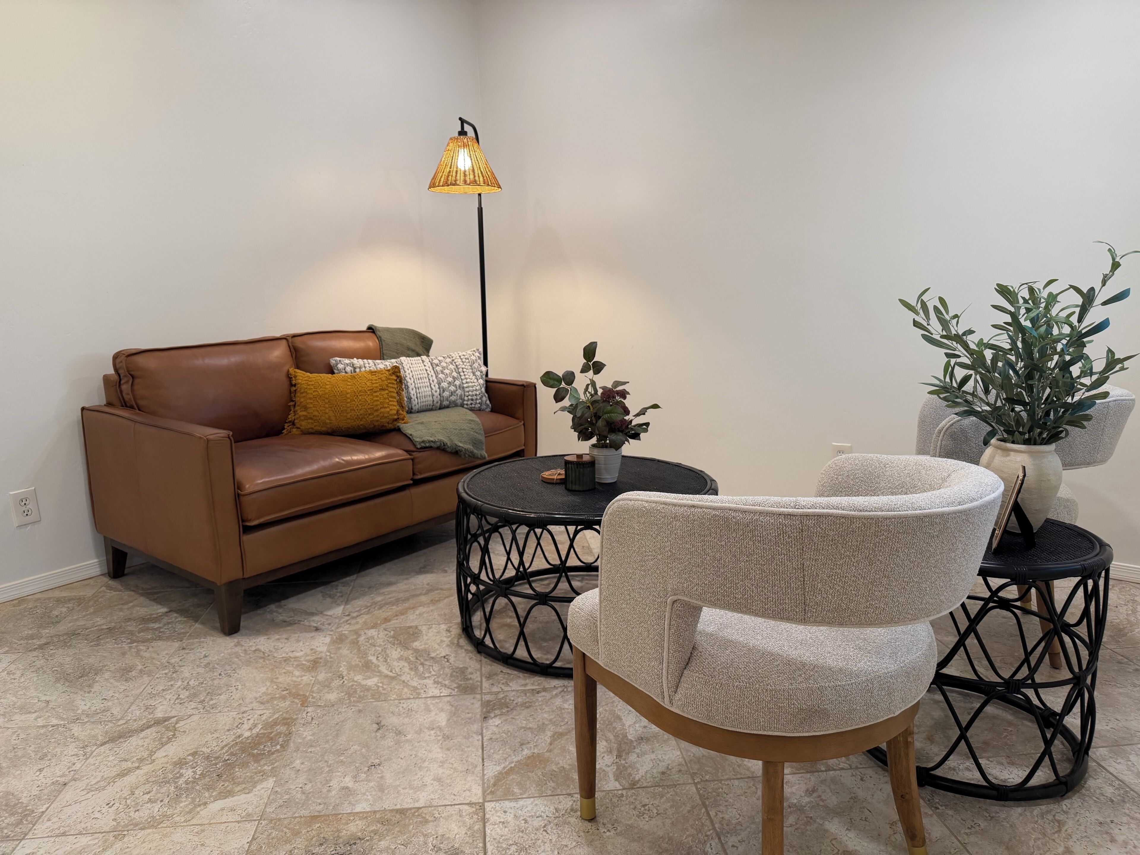 A living room with a brown leather sofa, a round coffee table, a patterned armchair, a floor lamp, and a potted plant.