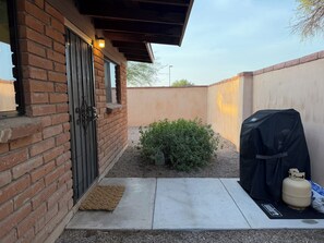 Un edificio in mattoni con una porta, un piccolo giardino con un arbusto, un barbecue e una bombola di propano.
