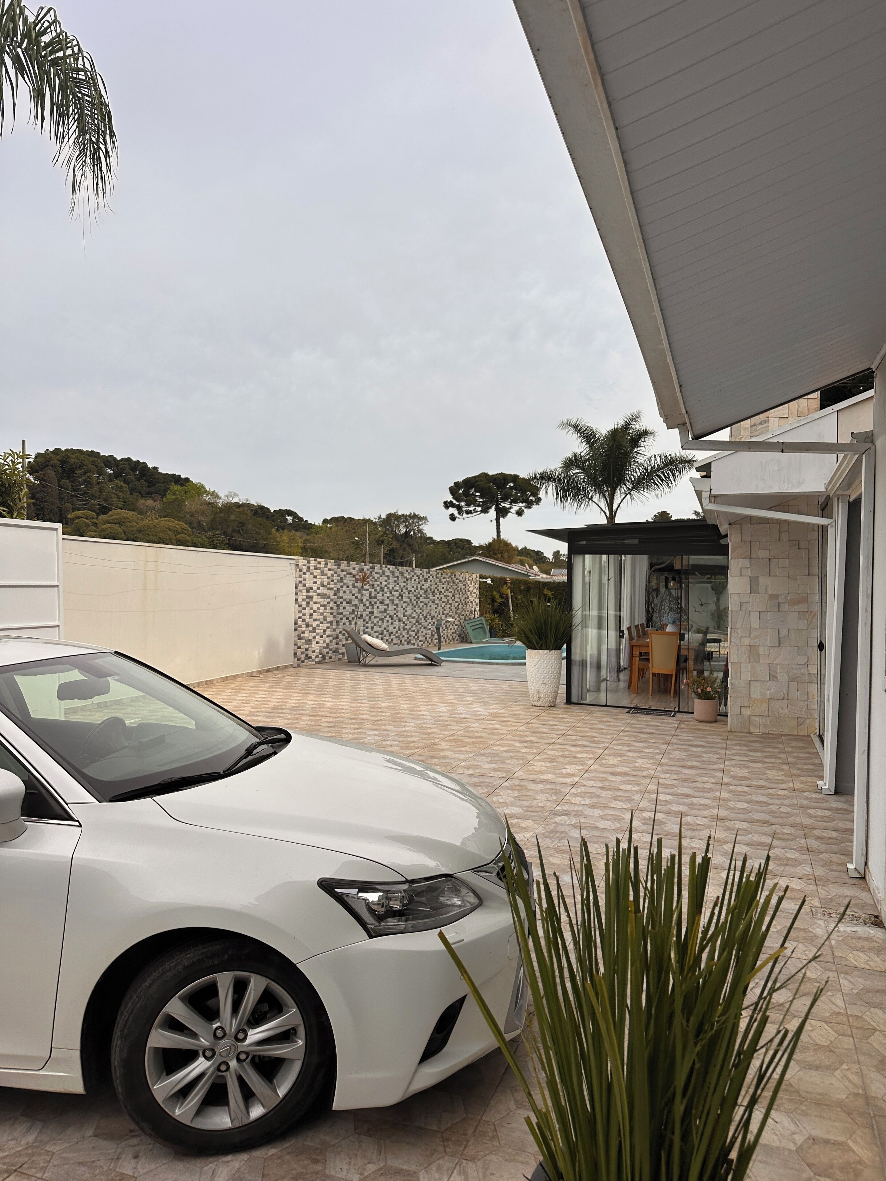 Um carro branco estacionado numa entrada de garagem com um espaço de pátio em mosaico, uma piscina e uma área coberta para estar ao ar livre.