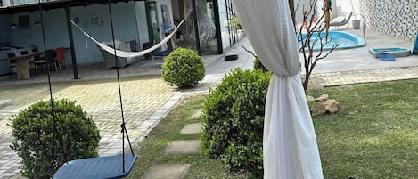 A covered outdoor area with a hammock, a swing, a pool, and a patio with a view of a building.