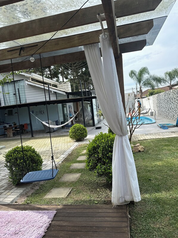 A covered outdoor area with a hammock, a swing, a pool, and a patio with a view of a building.
