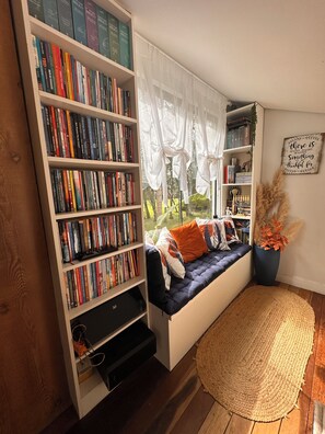A cozy reading nook with a built-in bookshelf, a cushioned bench, and a view through sheer curtains.
