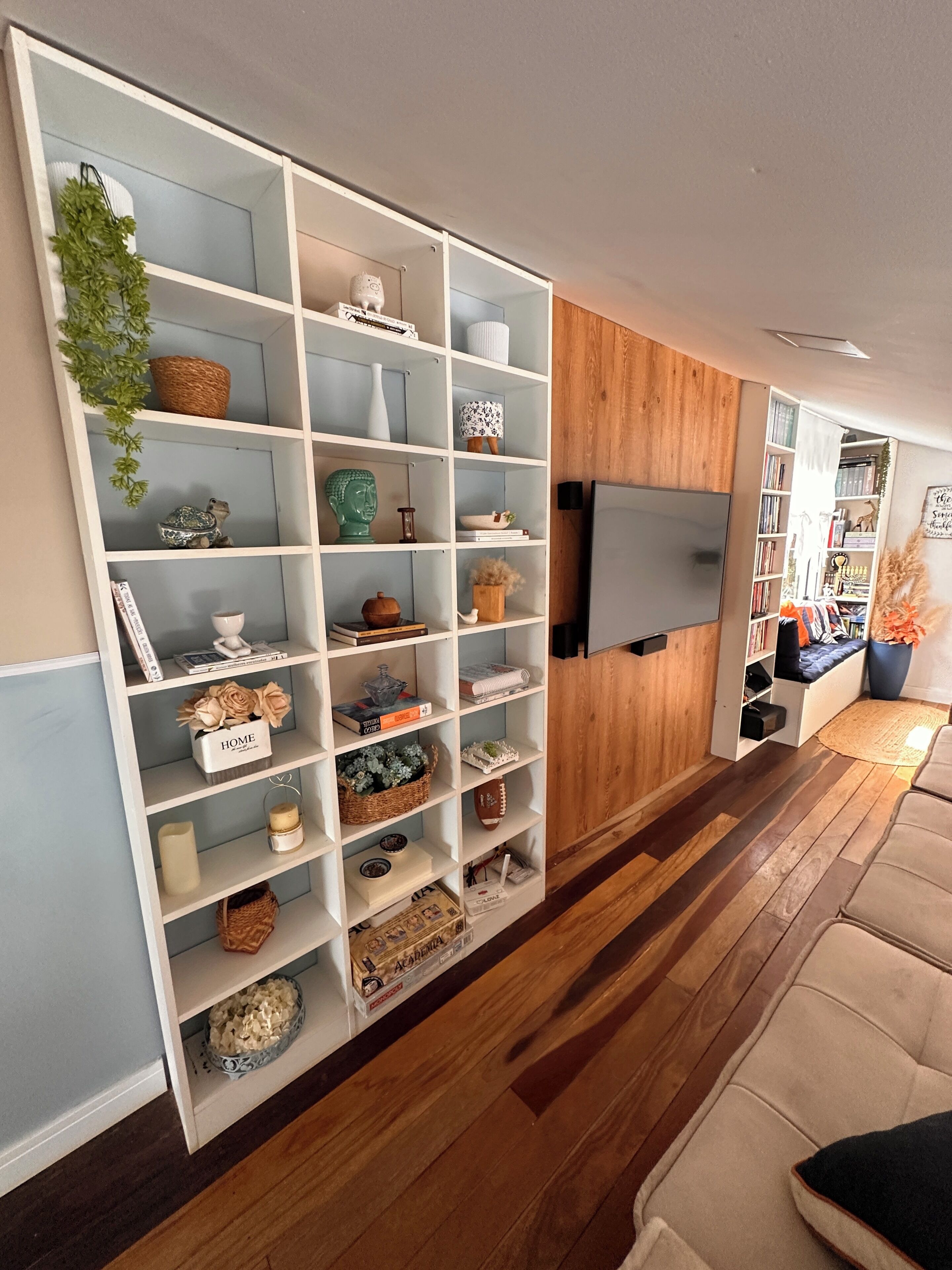 Una sala de estar con una estantería blanca llena de objetos decorativos, un televisor de pantalla plana en la pared y piso de madera.