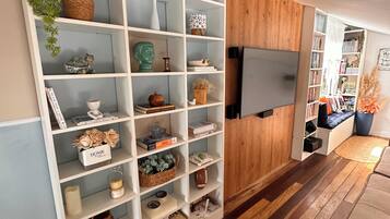 Una sala de estar con una estantería blanca llena de objetos decorativos, un televisor de pantalla plana en la pared y piso de madera.