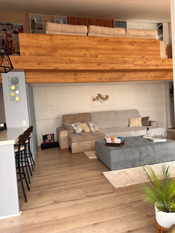 A loft-style living space with a high ceiling, a sofa, a coffee table, and a kitchen area with bar stools.