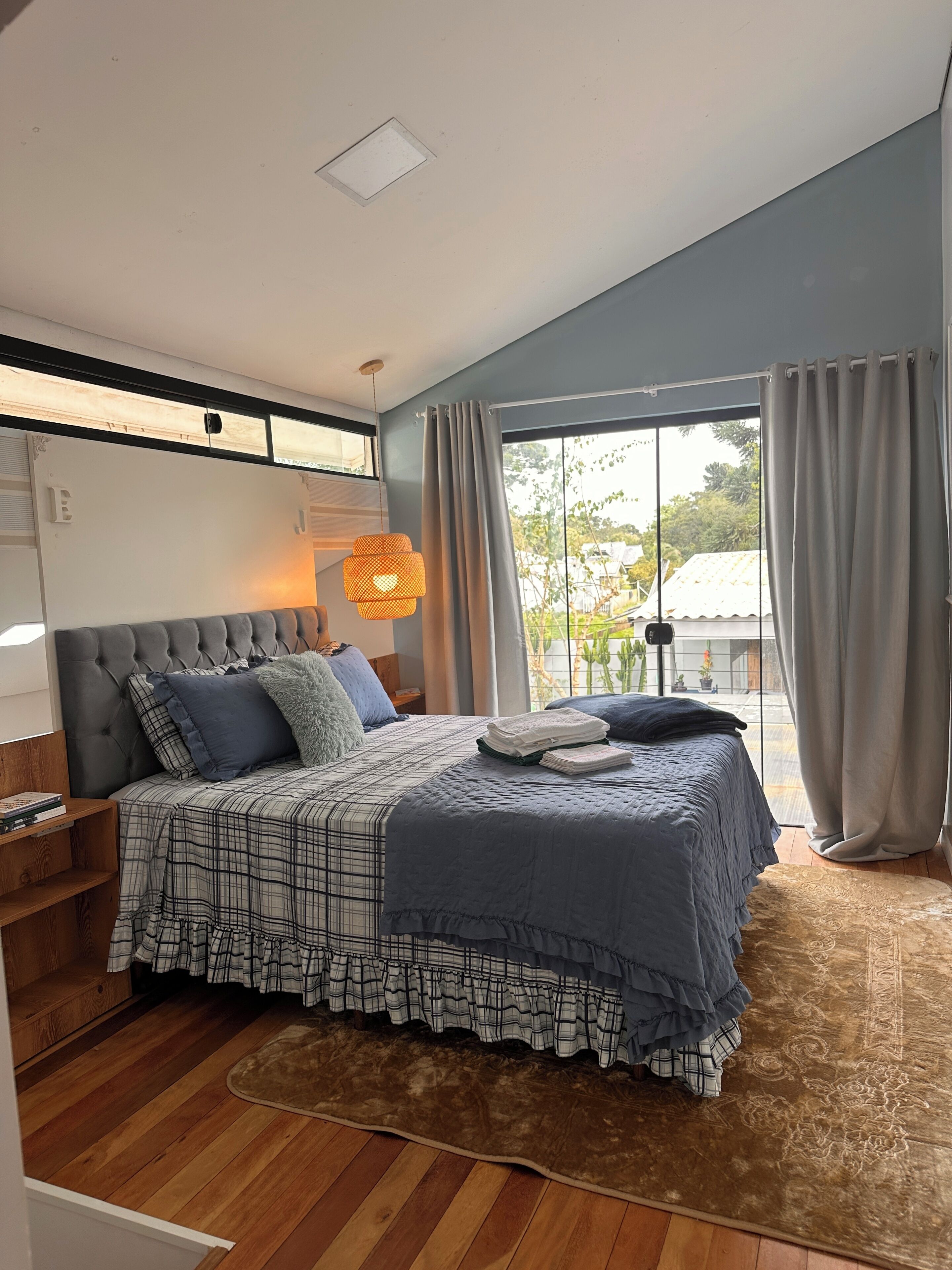 A bedroom with a large bed, a window with curtains, a wooden floor, and a ceiling light.