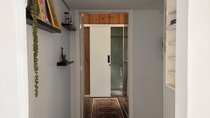 A hallway with a wooden floor, a white door, and a glass door leading to a bathroom. There are shelves with decorative items and a hanging plant.