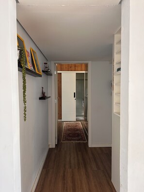 A hallway with a wooden floor, a white door, and a glass door leading to a bathroom. There are shelves with decorative items and a hanging plant.