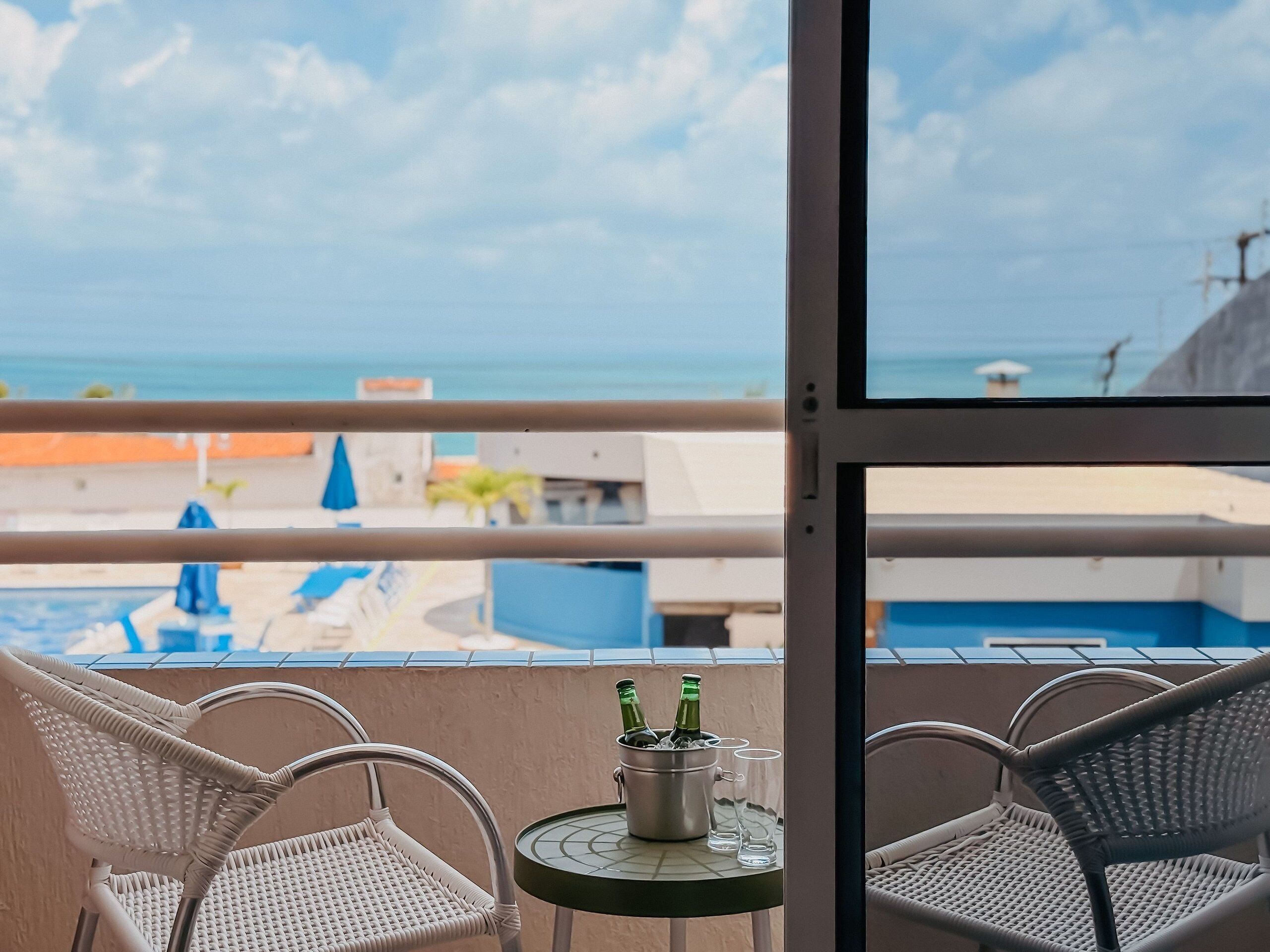 Un balcone con sedie in vimini, un tavolino con bottiglie e bicchieri, e vista su piscina e spiaggia.
