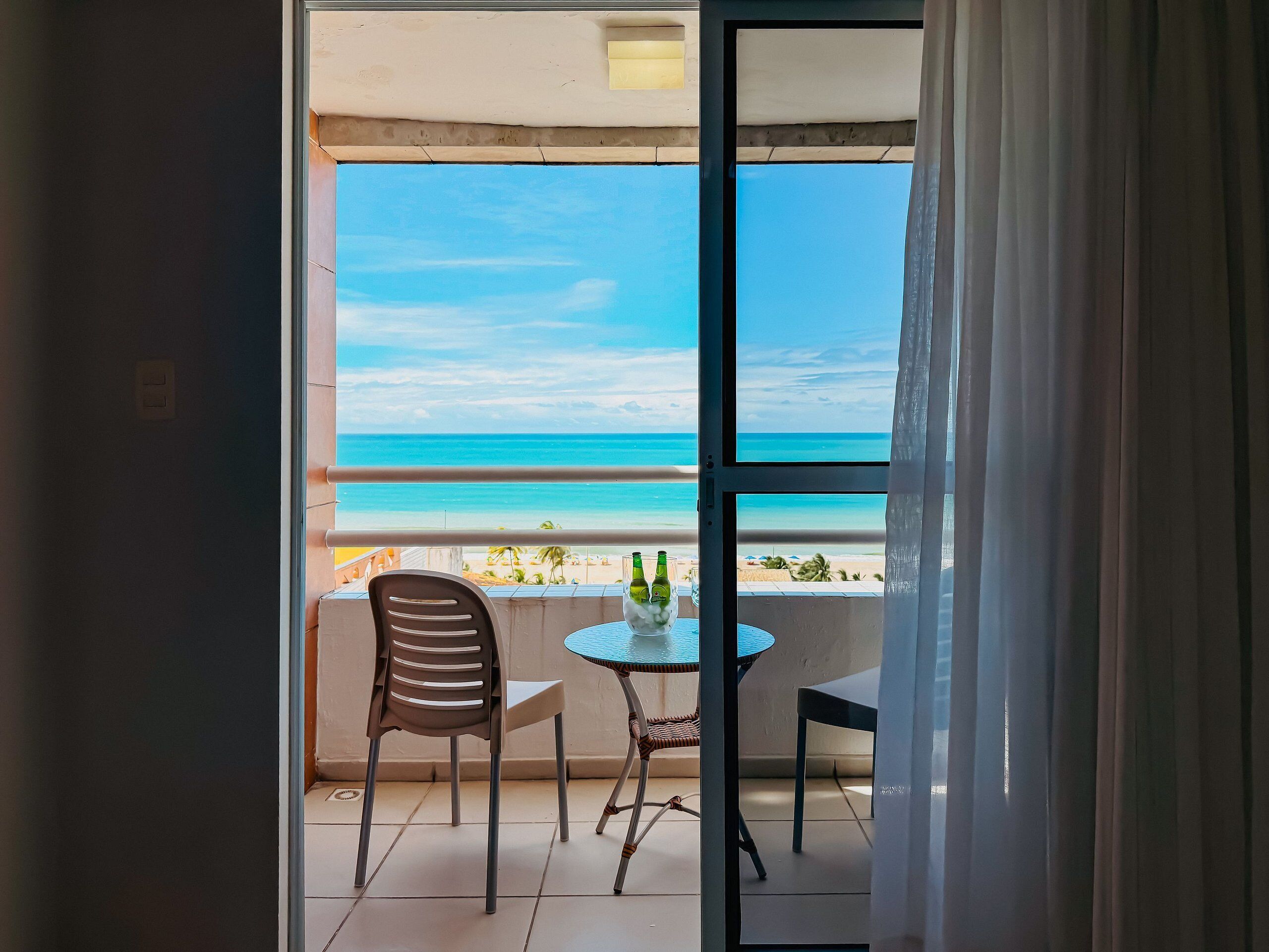 A balcony with a view of the beach, a table with bottles, and two chairs.