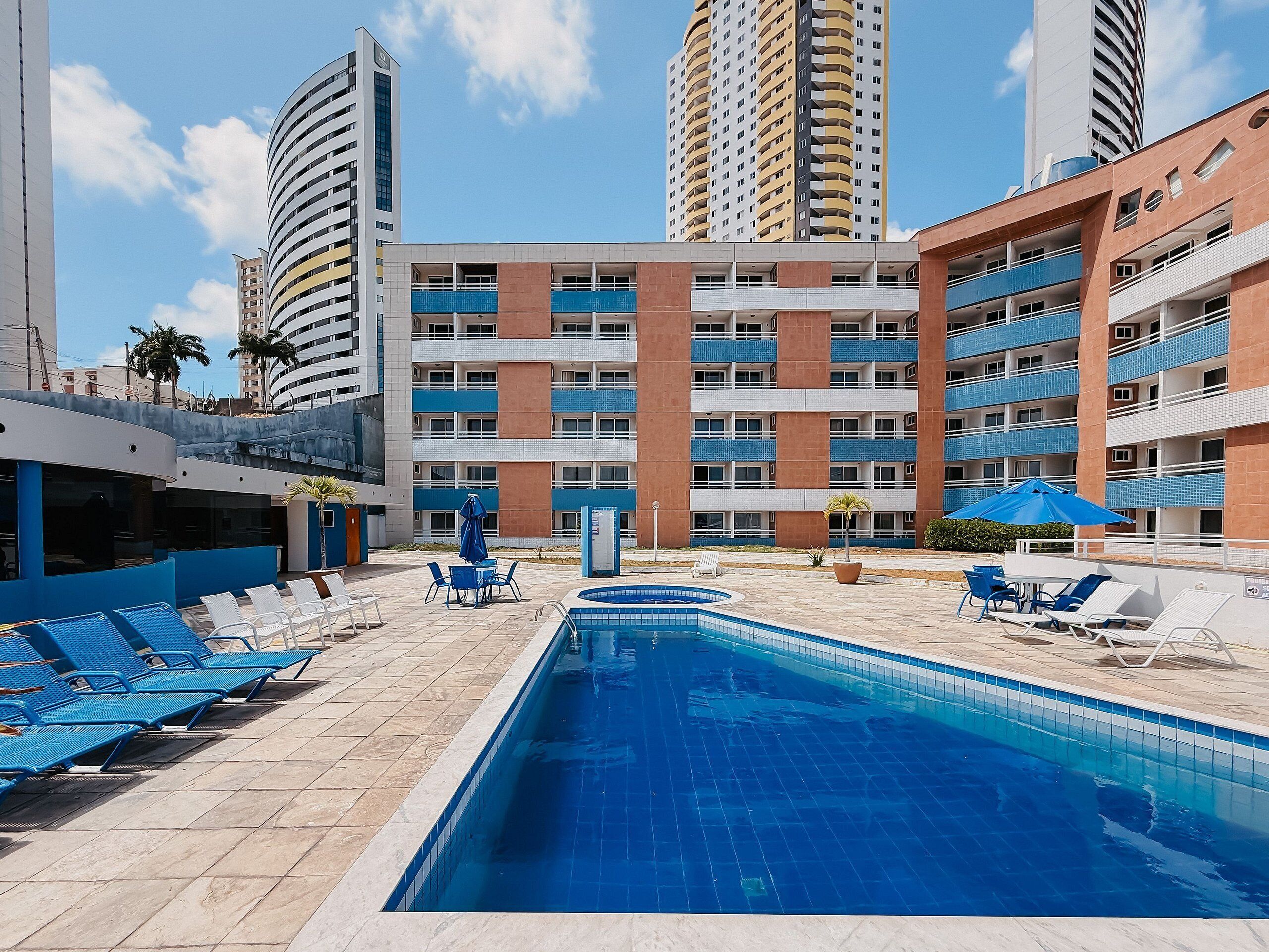 Un área de piscina de hotel con sillas azules, una piscina y un edificio con varios balcones.