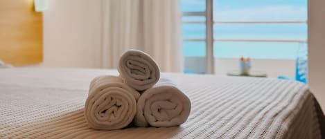 A neatly made bed with rolled white towels and a view of the sky through a window.