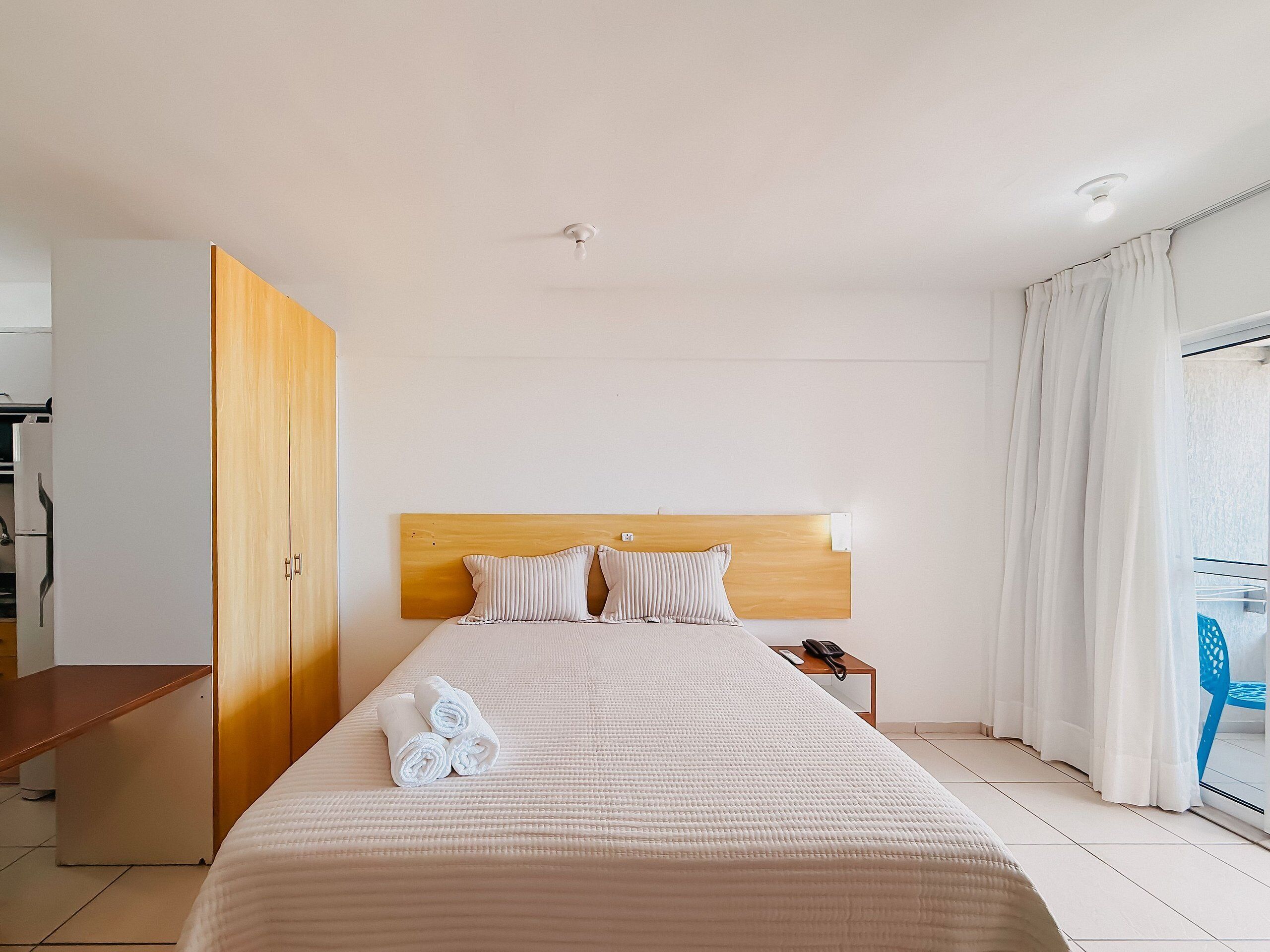 Une chambre d’hôtel avec un lit, une armoire en bois, une table de chevet avec un téléphone et une vue par la fenêtre, qui est dotée de rideaux.