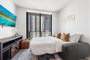 Ein modernes Schlafzimmer mit einem Bett, einem Fernseher, einem Fenster mit Blick auf die Stadt und einem Wanddekor.