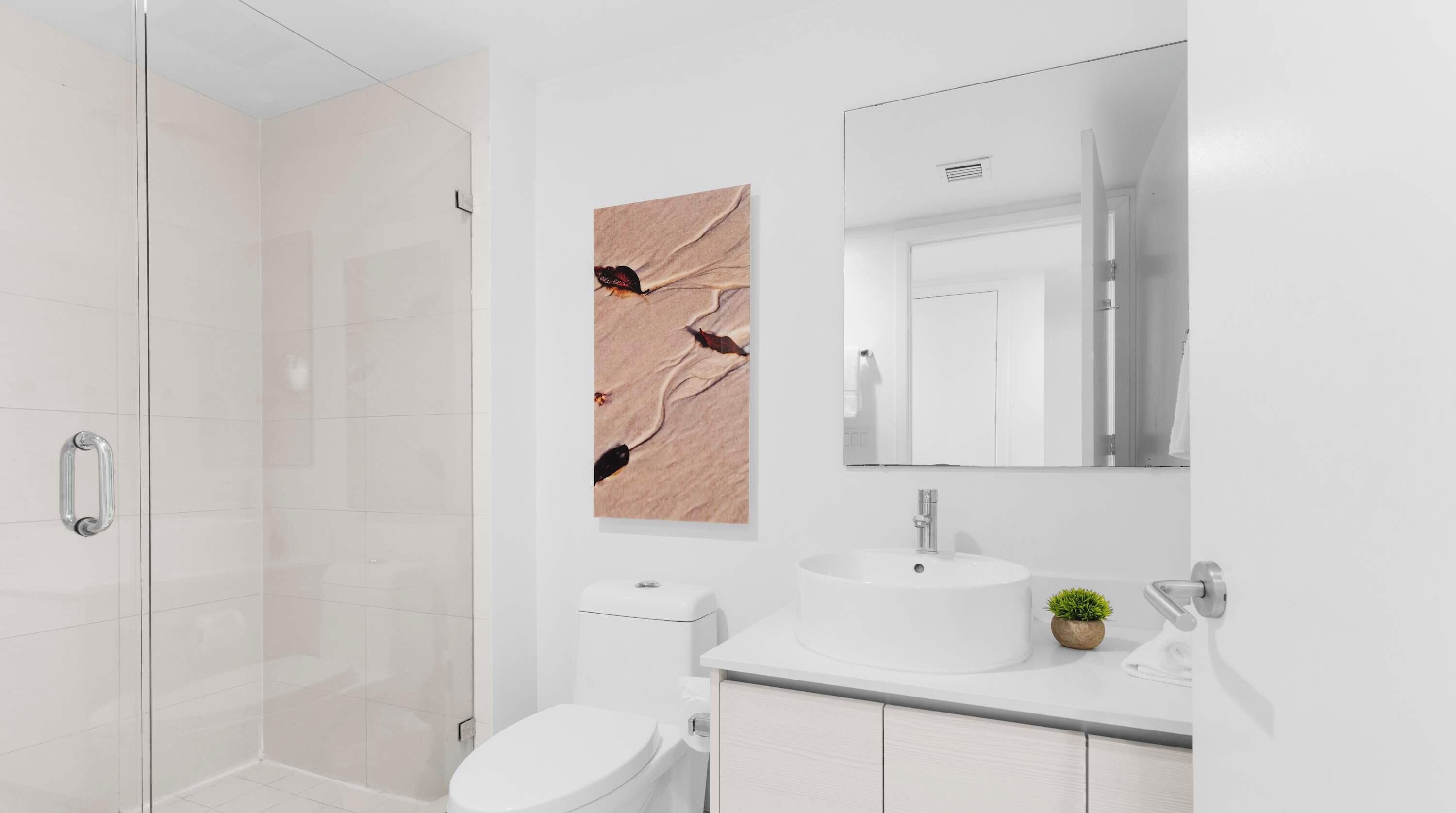 Ein modernes Badezimmer mit Duschkabine aus Glas, einer weißen Toilette, einem Waschbecken mit Spiegel darüber und einem an der Wand hängenden Kunstwerk.