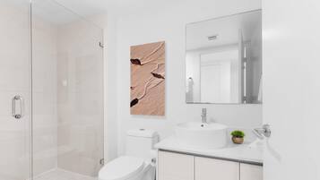 Ein modernes Badezimmer mit Duschkabine aus Glas, einer weißen Toilette, einem Waschbecken mit Spiegel darüber und einem an der Wand hängenden Kunstwerk.