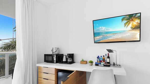 A hotel room with a white desk, a flat-screen TV displaying a beach scene, a coffee maker, a microwave, and a small plant. - Modern Studio Resort Style w/ Private Balcony (Hallandale Beach)