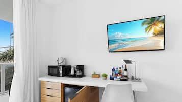Ein Hotelzimmer mit weißem Schreibtisch, einem Flachbildfernseher, der eine Strandansicht zeigt, einer Kaffeemaschine, einer Mikrowelle und einer kleinen Pflanze.