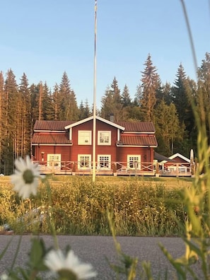 A red wooden building with a white fence in front, surrounded by tall trees.