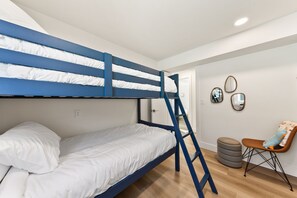 Un lit superposé avec une échelle, une chaise et un petit pouf dans une pièce au plancher en bois et aux murs blancs.