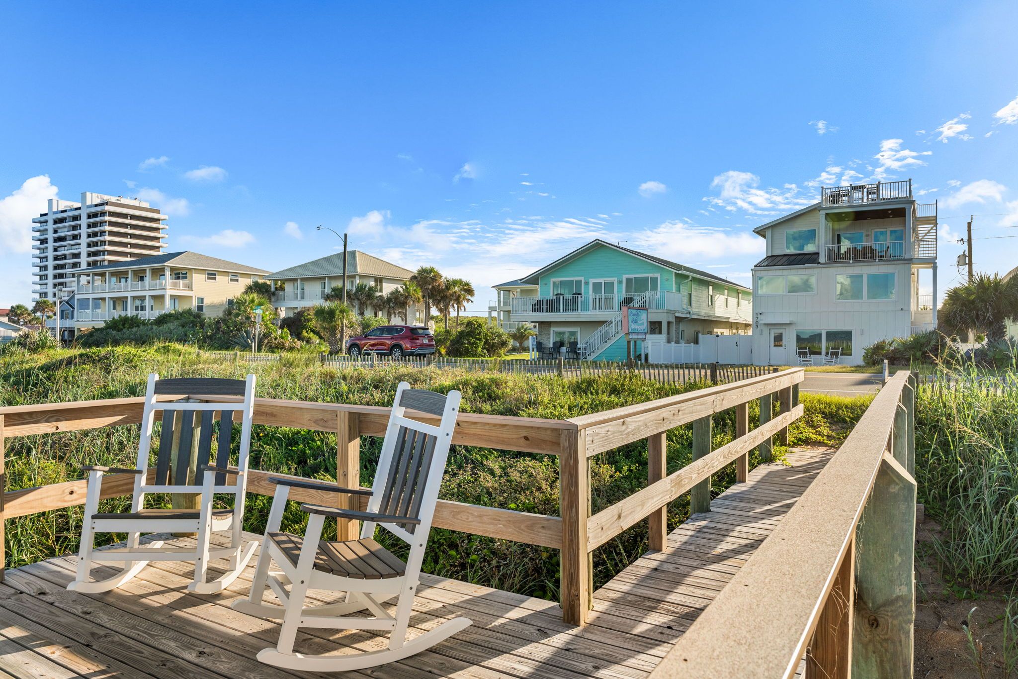 A wooden deck with two rocking chairs, a wooden fence, and a view of beachfront buildings.