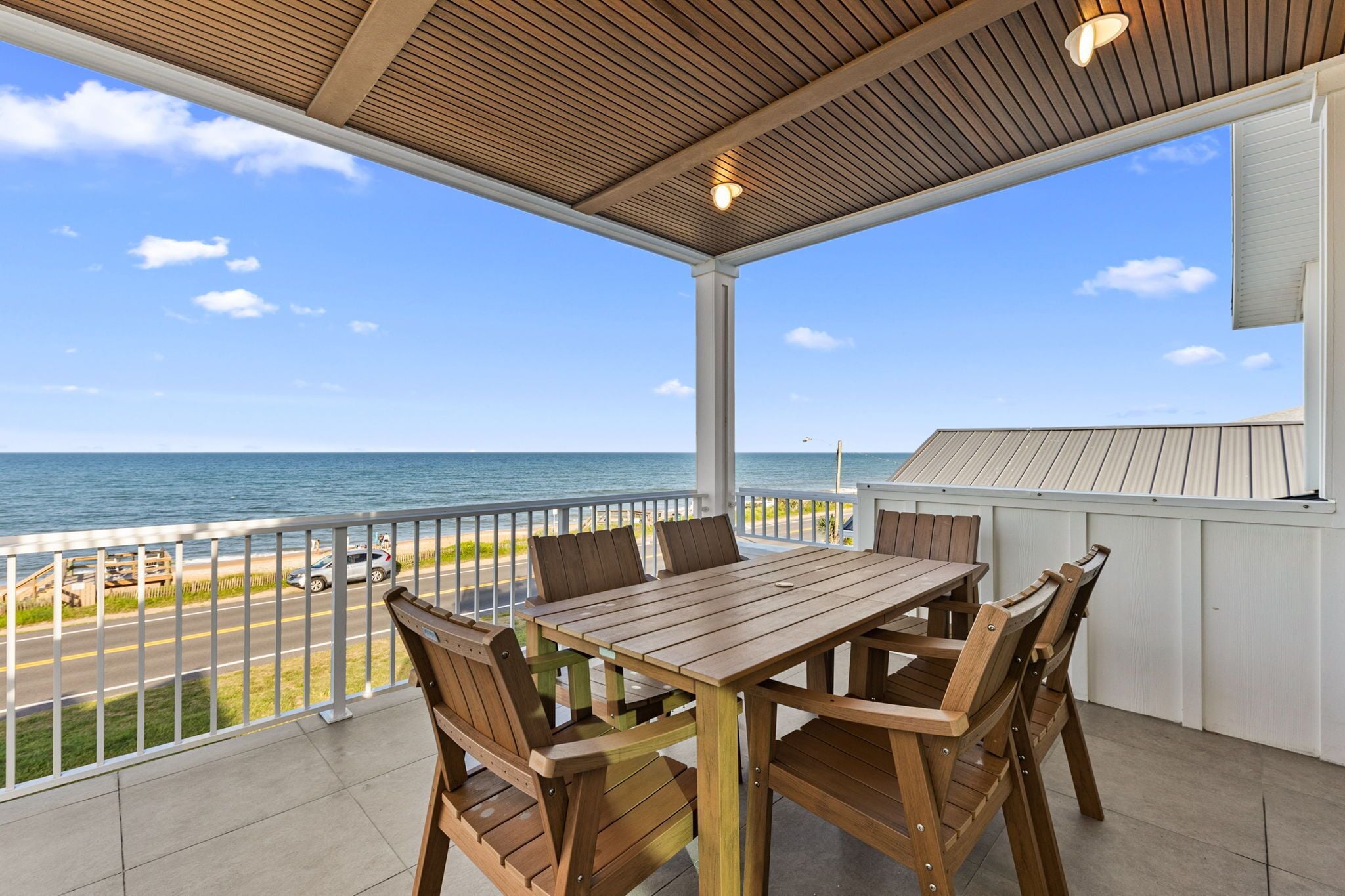 A covered patio with a wooden table and chairs, overlooking a beach and road.