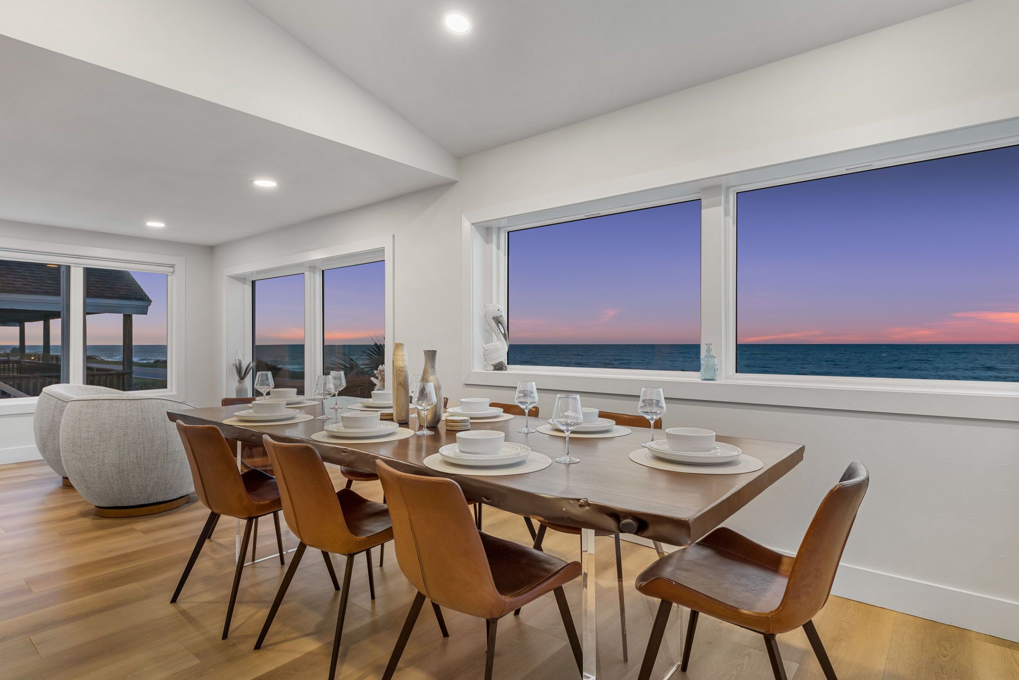 Un comedor con vista al océano al atardecer, con una mesa larga y sillas, y un amplio ventanal.