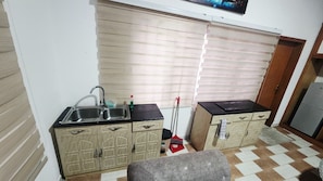 A compact kitchen with a sink, two cabinets, a broom, and a window with blinds. - 1 BR Cozy Furnished Apartment With Private bath & Premium Amenities (Accra)
