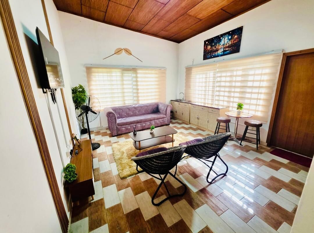 Uma sala de estar com um sofá, uma mesa de centro e um espaço de jantar com cadeiras. Uma cozinha pode ser vista ao fundo.