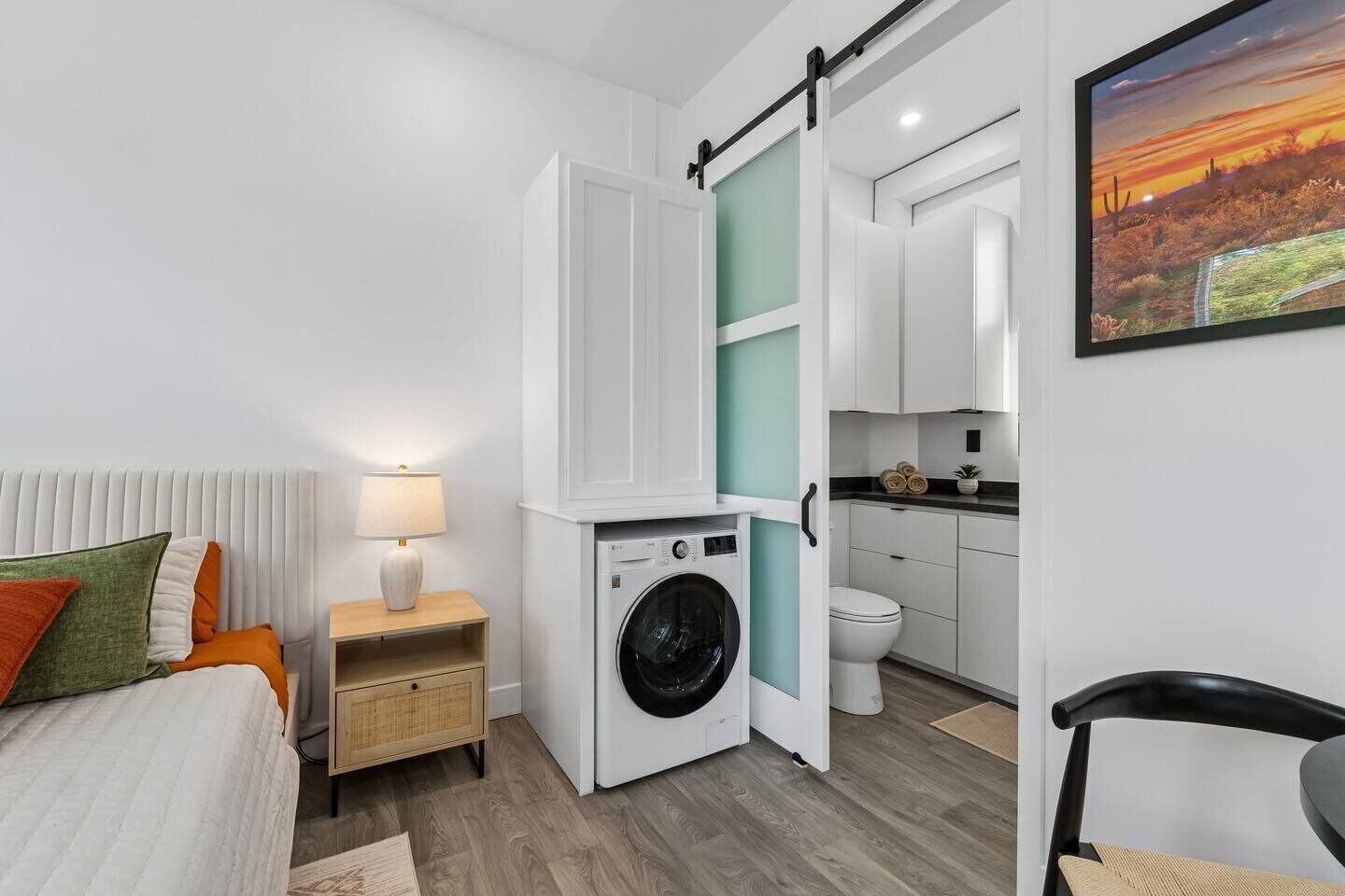 A compact hotel room with a bed, a bedside table with a lamp, a washing machine, a kitchenette, and a bathroom.