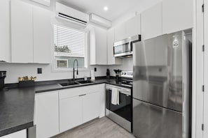 A modern kitchen with white cabinetry, a stainless steel refrigerator, a built-in oven, and a window with blinds. - Modern Boxabl Casita | Stillwater OK | Pasadera 4 (Stillwater)
